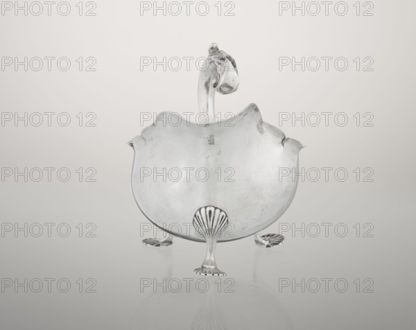 Sauce boat by Henry Matthews fl. 1893–ca. 1930, goldsmith. Hemispherical silver sauce boat with an elongated wide spout and irregularly cut rim, supported by three palmette feet; profiled volute handle decorated with an acanthus leaf. Dated 1903–1904, Birmingham, England. Vessel. Height 11.5 cm, width 16 cm, depth 9.7 cm, weight 248 g., A reflective silver object shaped like a shallow bowl with a wavy rim sits on a smooth surface, supported by three ornate feet resembling shells and small flared bases; a curved handle rises from the back of the bowl and ends in a rounded decorative knot. The scene shows tones of silver, gray, white, and subtle off-white in the background and surface, with soft reflections of the object visible beneath it.