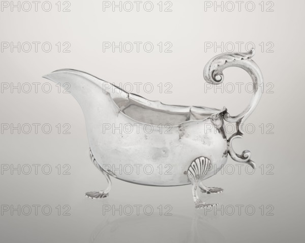 Sauce boat, Henry Matthews fl. 1893–ca. 1930, goldsmith. Hemispherical silver sauce boat with an elongated wide spout and an irregularly cut rim; supported by three palmette feet; volute handle, profiled and decorated with an acanthus leaf. 1903–1904. Birmingham, England. Vessel. Height 11.5 cm; width 16 cm; depth 9.7 cm; weight 248 g., A polished silver gravy boat with an ornate curled handle and a curved pouring spout rests on a reflective surface; the vessel stands on a few decorative feet shaped like shell motifs and small clawed pads, and its body shows smooth rounded contours with subtle dents and surface marks; the background is a gradient of light gray to off-white, and visible colors include silver, gray, off-white, and a hint of warm beige in the reflection.