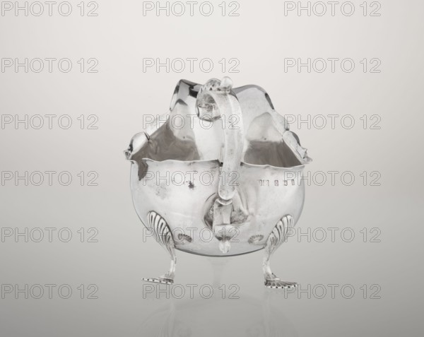 Sauce boat; maker: Henry Matthews fl. 1893–ca. 1930, goldsmith; hemispherical silver sauce boat with an elongated wide spout and irregularly cut rim; supported by three palmette feet; volute handle profiled and decorated with an acanthus leaf; ornament: acanthus, volutes, palmettes; date: 1903–1904; place: Birmingham, England, Great Britain; vessel; height 11.5 cm, width 16 cm, depth 9.7 cm; weight 248 g., A reflective metal bowl with a scalloped rim sits on a smooth surface, supported by a group of slender, curved legs ending in flat, decorative feet; a thick, curved handle arches over the top and attaches to the body with rounded fittings, and the bowl's interior shows smooth, shiny metal with subtle surface marks and reflections. The object displays metallic silver tones with areas of darker gray patina and slight shadowing, set against a soft, light gray to off-white gradient background.