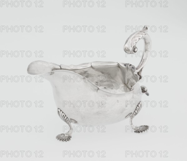 Sauce boat, by Henry Matthews fl. 1893–ca. 1930, goldsmith; hemispherical silver sauce boat with an elongated wide spout and irregularly cut rim, supported by three palmette feet; volute handle, profiled and decorated with an acanthus leaf; decorated with acanthus, volutes, and palmettes; dated 1903–1904; Birmingham, England, Great Britain; vessel; height 11.5 cm, width 16 cm, depth 9.7 cm, weight 248 g., A silver-colored metal pitcher with a curved spout and an ornate scrolling handle sits on a plain light-gray background; the pitcher has a rounded body with a wavy rim and a slightly dimpled interior showing reflective highlights, and it stands on a group of decorative claw-like feet with shell-shaped bases that share the same silver color.