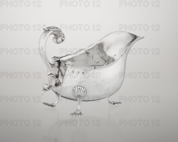 Sauce boat; maker: Henry Matthews fl. 1893–ca. 1930, goldsmith; hemispherical silver sauce boat with an elongated wide spout and irregularly cut rim; supported by three palmette feet; volute handle, profiled and decorated with an acanthus leaf; ornament: acanthus, volutes, palmettes; date: 1903–1904; place: Birmingham, England, Great Britain; vessel; height 11.5 cm, width 16 cm, depth 9.7 cm, weight 248 g, A polished metal vessel with a curved spout and an ornate scrolling handle sits on three decorative feet; the vessel is reflective and shows soft distorted reflections, with smooth and slightly textured surfaces. The handle and feet feature curved, shell-like and leaf-like motifs, and the interior of the vessel has reflective ripples. The overall colors visible are silver tones, with gradients of gray and subtle white highlights against a pale gray background.