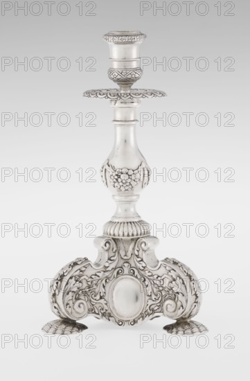 Single-candle candlestick, neo-Baroque style, silver, supported on three shells and a three-sided raised base with side edges composed of volutes, richly decorated with floral tendril and plain medallions; baluster shaft at the bottom enclosed in a godron half-cylinder, decorated with floral festoons above, topped with a plate-shaped profiled drip pan with a cylindrical sleeve. Maker: Martin Goldstein fl. 1878–1886 and the firm C.J. Vander Ltd., London. Dated 1884/1885. Function: lighting. Dimensions: height 24 cm; width 13.6 cm; depth 13.6 cm; weight 852 g. Location: London, England Great Britain., The image shows a single ornate metal candlestick with a detailed decorative stem and base, featuring floral and scroll motifs; the metal appears predominantly silver with darker tarnished accents highlighting the engraved patterns, and the background is a gradient of light gray to white.