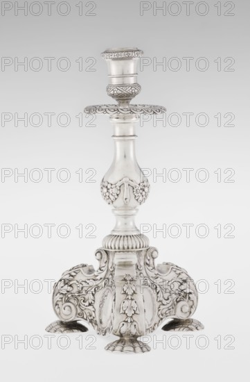 Single-candle candlestick, maker: Martin Goldstein fl. 1878–1886; maker firm: C.J. Vander, Ltd. London; goldsmith's firm, est. 1856; provenance: purchase scrap - purchase. Neo-Baroque style. Ornament: medallion, volutes, floral ornaments, godron ornaments, floral tendril. Description: single-candle candlestick in silver, supported on three shells and a three-sided raised base with side edges composed of volutes; richly decorated with floral tendril and plain medallions; baluster shaft at the bottom enclosed in a godron half-cylinder, decorated with floral festoons above, topped with a plate-shaped profit with a cylindrical sleeve. Date: 1884/1885. Place: London, England, Great Britain. Function: lighting. Dimensions: height 24 cm; width 13.6 cm; depth 13.6 cm; weight 852 g., A single ornate metallic candlestick stands upright against a light gray background, finished in a bright silver tone with darker areas where detailing creates contrast; the piece features a rounded top socket with...