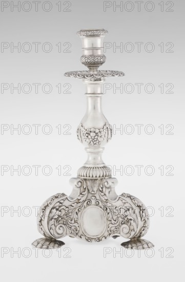 Single-candle candlestick by Martin Goldstein fl. 1878–1886 and the firm Vander, C.J., Ltd. London; goldsmith's firm, est. 1856. Neo-Baroque style; silver. Supported on three shells and a three-sided raised base with side edges composed of volutes, richly decorated with floral tendrils and plain medallions. Baluster shaft with bottom enclosed in a godron half-cylinder, decorated with floral festoons above, topped with a plate-shaped profiled element and a cylindrical sleeve. Ornamentation includes medallion, volutes, floral and godron ornaments. Dated 1884/1885, London, England. Function: lighting. Height 24 cm; width 13.6 cm; depth 13.6 cm; weight 852 g., A single ornate candlestick stands against a plain light background, made of a shiny metallic material with silver and gray tones; the candlestick has a flared holder at the top with decorative banding and a scalloped drip plate beneath it, a smooth tapered central column that transitions into a rounded midsection embellished with ra...