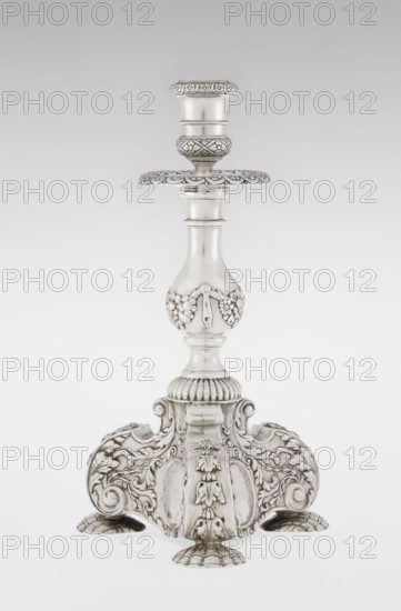 Single-candle candlestick by Martin Goldstein fl. 1878–1886 and Vander, C.J., Ltd. London; goldsmith's firm, est. 1856. Neo-Baroque style; silver. Supported on three shells and a three-sided raised base with side edges composed of volutes; richly decorated with floral tendrils and plain medallions. Baluster shaft at the bottom enclosed in a godron half-cylinder, decorated with floral festoons above, topped with a plate-shaped profiled element with a cylindrical sleeve. Medallion, volutes, floral and godron ornaments. Dated 1884/1885, London, England. Function: lighting. Height 24 cm; width 13.6 cm; depth 13.6 cm; weight 852 g., A single ornate metal candlestick stands upright against a neutral pale background, showing a polished silver color with darker patina in recesses; the stem tapers and swells with smooth rounded sections and a bulbous midsection decorated with raised floral garlands and ribbon-like motifs, a scalloped round drip pan with engraved petal patterns sits near the top...