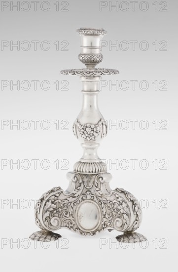 Single-candle candlestick by Martin Goldstein fl. 1878–1886 and C.J. Vander Ltd. London; goldsmith's firm, est. 1856. Neo-Baroque style; silver. Supported on three shells and a three-sided raised base with side edges composed of volutes; richly decorated with floral tendrils and plain medallions. Baluster shaft at the bottom enclosed in a godron half-cylinder, decorated with floral festoons above; topped with a plate-shaped profiled drip pan with a cylindrical sleeve. Medallion, volutes, floral and godron ornaments. Date: 1884/1885. Place: London, England, Great Britain. Function: lighting. Dimensions: height 24 cm; width 13.6 cm; depth 13.6 cm. Weight: 852 g., A single ornate metal candlestick stands upright with a polished silvery surface, featuring a flared top, a decorative drip plate with scalloped edges, and a baluster stem embellished with raised floral motifs; the base broadens into an elaborately carved tripod platform with scrolling acanthus leaves, shell-like feet, and an ov...