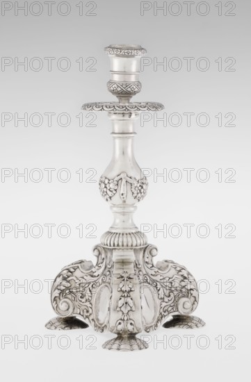 Single-candle candlestick by Martin Goldstein fl. 1878–1886 and Vander, C.J., Ltd. London; goldsmith's firm, est. 1856. Neo-Baroque style; silver. Supported on three shells and a three-sided raised base with side edges composed of volutes, richly decorated with floral tendrils and plain medallions. Baluster shaft at the bottom enclosed in a godron half-cylinder, decorated with floral festoons above, topped with a plate-shaped drip pan and a cylindrical sleeve. Medallion, volutes, floral and godron ornaments. Date: 1884/1885. Place: London, England, Great Britain. Function: lighting. Dimensions: height 24 cm; width 13.6 cm; depth 13.6 cm. Weight: 852 g., A single ornate metal candlestick stands against a plain light background, showing a polished silver surface with darker toned recesses that highlight intricate carving and relief work; the base flares into a decorative tri-lobed platform with a mix of scrolls, acanthus-like leaves, shell-shaped feet, and raised floral motifs, while the...