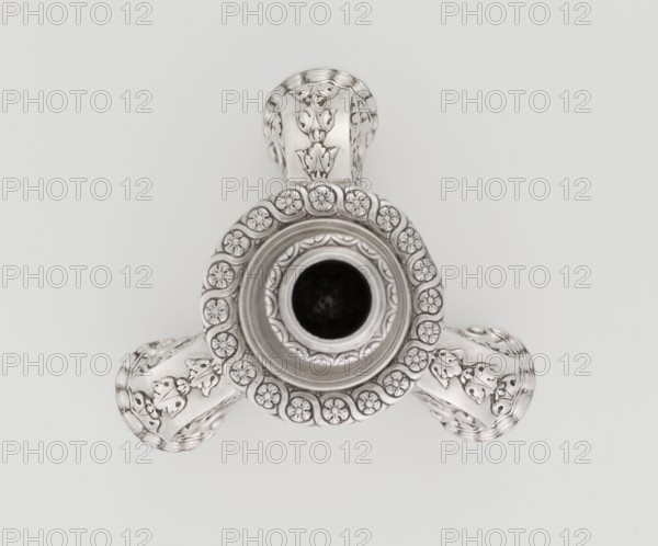 Single-candle candlestick by Martin Goldstein fl. 1878–1886 and the firm Vander, C.J., Ltd. London; goldsmith's firm, est. 1856. Neo-Baroque style, 1884/1885, England London, Great Britain. Silver; supported on three shells and a three-sided raised base with side edges composed of volutes, richly decorated with floral tendril and plain medallions. Baluster shaft at the bottom enclosed in a godron half-cylinder, decorated with floral festoons above, topped with a plate-shaped knop and a cylindrical sleeve. Ornament: medallion, volutes, floral ornaments, godron ornaments, floral tendril. Function: lighting. Height 24 cm; width 13.6 cm; depth 13.6 cm; weight 852 g., A top-down view of a metallic object with a central circular opening surrounded by a decorative rim featuring a ring of flower-like motifs, and three ornate curved arms extending outward; the surface shows embossed floral and leaf patterns and a polished reflective finish. The visible colors are silver and shades of gray again...