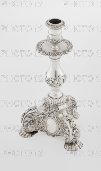 Single-candle candlestick, neo-Baroque style, silver, supported on three shells and a three-sided raised base with side edges composed of volutes, richly decorated with floral tendril and plain medallions; baluster shaft at the bottom enclosed in a godron half-cylinder, decorated with floral festoons above, topped with a plate-shaped profit, with a cylindrical sleeve. Medallion, volutes, floral ornaments, godron ornaments, floral tendril. Date: 1884–1885, London, England Great Britain. Function: lighting. Dimensions: height 24 cm; width 13.6 cm; depth 13.6 cm; weight 852 g., A silver-colored ornate candlestick with a decorative circular cup at the top, a flared scalloped drip tray beneath it, and a turned column that narrows then widens into a bulbous midsection adorned with raised floral garlands; the base is an elaborately carved three-footed platform with scrolling acanthus motifs, shell-like feet, and oval cartouche panels, all rendered in metallic silver and subtle darker patina a...