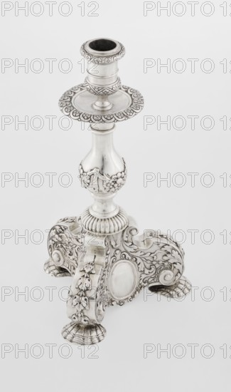 Single-candle candlestick, neo-Baroque style, silver, 1884/1885, England, London, Great Britain. Supported on three shells and a three-sided raised base with side edges composed of volutes, richly decorated with floral tendril and plain medallions. Baluster shaft at the bottom enclosed in a godron half-cylinder, decorated with floral festoons above, topped with a plate-shaped profit with a cylindrical sleeve. Medallion, volutes, floral ornaments, godron ornaments, floral tendril. Height 24 cm, width 13.6 cm, depth 13.6 cm, weight 852 g. Makers: Martin Goldstein fl. 1878–1886 and Vander C.J., Ltd London; goldsmith's firm, 1856–. Function: lighting., A silver-colored ornate candlestick with a decorative top rim and a shallow dish beneath the holder, a baluster-shaped stem adorned with floral relief, and a heavily ornamented tripod base with scrolling patterns, leaf motifs, rounded medallion areas, and shell-like feet, all shown against a plain light-gray background.