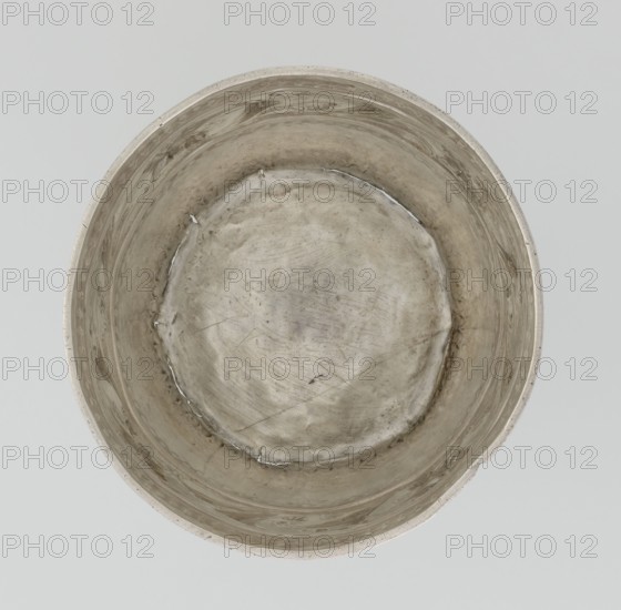 Bowl by William I Sumner fl. 1773–ca. 1807, goldsmith. Circular, bulging vessel with a flat bottom and flared upper edge; decorated on the belly with a wide band of relief ornament of acanthus leaves and flowers. Materials: metal vessel. Styles: Georgian, George III, Empire; motifs: acanthus. Date: 1804–1805. Place: London, England. Dimensions: height 8 cm; diameter 11.5 cm; weight 145 g. Provenance unknown., A top-down view of a round metal container with a dull silver-gray surface showing irregular scratches, scuffs, and areas of darker tarnish; the interior rim and walls display subtle swirling patterns and a few small raised seams, and the bottom appears slightly concave with uneven texture and faint shadowing; the background is a flat pale gray.