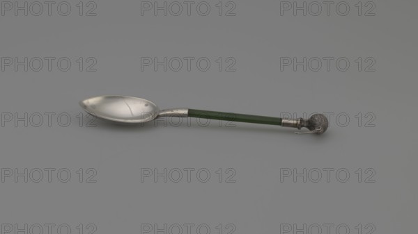 Souvenir spoon by FH goldsmith; Birmingham; fl. ca. 1903–1904. Souvenir spoon with an oval scoop, a flat jade handle set in a silver sleeve, and topped with a silver full-plastic kiwi bird figurine. Likely made in Birmingham, England, ca. 1903–1904. Cutlery; height 10.8 cm, width 2.3 cm., A single spoon lies horizontally on a plain background, showing a metallic bowl with a smooth, slightly reflective surface and some small marks, a straight handle that is green and cylindrical, and a decorative metallic end shaped like a textured sphere with a small protruding element; the visible colors include silver, green, and shades of gray in the background.