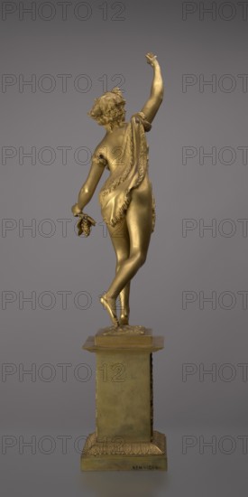 Candlestick, lyre and swans iconography, 19th century, Paris, France, lighting, height 54 cm, A gilded statue of a female figure standing on a square pedestal, shown from the back with one arm raised and the other lowered holding a small cluster of objects; the figure wears a draped cloth over one shoulder and around the waist with visible fringed edges, and has curly hair adorned with small decorative elements; the pedestal and statue share a metallic gold color, and the background is a uniform muted gray.