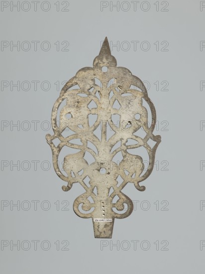 Hinge end for a chest; fittings, hinge end from a pair, in the form of an openwork rounded plaque with a tendril motif, two male masks among the twists. Hinge cut off; perhaps originally a disc hinge with a sleeve at the end of the plate or a strap hinge in the form of a clock hand. 17th century, Poland, Central Europe. Door fittings. Height 21.5 cm, width 12.5 cm, depth 0.2 cm. Maker: Roman Szewczykowski 1849–1901, An ornate, flat metal object with a symmetrical, openwork design featuring curving, scroll-like shapes and floral or leaf-like cutouts, showing a weathered surface with patches of darker and lighter areas; the metal appears mainly gray with areas of brownish tarnish and pale off-white spots, set against a uniform light gray background.