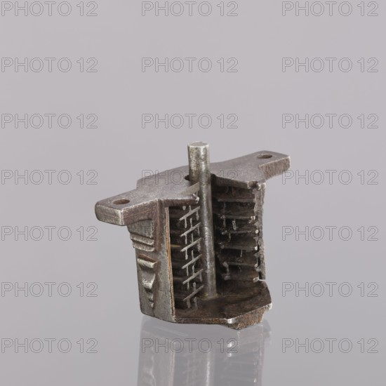 Castle register, unknown author; single-sided register with a key pin; octagonal plates; bottom extensions on the sides with screw holes; triangular profiled reinforcements on the walls; rectangular reinforcing strip on the top plate. Key with a tubular smooth shaft, with a bow/feather with seven vertical ribs, two recesses on one side and one on the other; extended, ending with an arched extension. 18th–19th century, Central Europe; metalwork; height 7.6 cm, width 9.9 cm, depth 5.1 cm., A small metal object sits against a pale gray background, showing a metallic dark gray and brownish surface with areas of lighter silver highlights; it has a vertical cylindrical post rising from a rectangular base that is partially hollowed to reveal a group of layered, ladder-like internal ribs, and the top extends into two flat horizontal wings with rounded holes near their ends; the metal surface appears textured and slightly worn, and the object casts a faint reflection on the glossy surface benea...