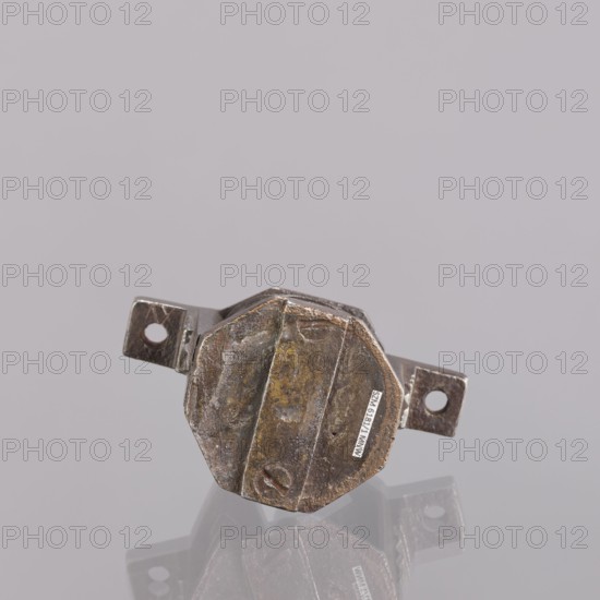 Castle register, gift, locksmithing. Single-sided register with a key pin, octagonal plates, bottom extensions on the sides with screw holes, triangular profiled reinforcements on the walls. A rectangular reinforcing strip on the top plate. Key with a tubular, smooth shaft, feather with seven vertical ribs with two recesses on one side and one on the other. Extended, ending with an arched extension. 18th–19th century, Central Europe, metalwork; height 7.6 cm, width 9.9 cm, depth 5.1 cm., A small metallic object with an octagonal central body and two flat protruding tabs, one on each side, sits on a reflective surface. The metal shows a worn, aged appearance with areas of brownish rust, dull goldy-brown patina, and darker gray tones. The surface has scratches, dents, and irregular textures, and a few shallow circular indentations are visible. The reflection of the object appears faintly beneath it, echoing the same colors and shapes. The background is a smooth, uniform pale gray.