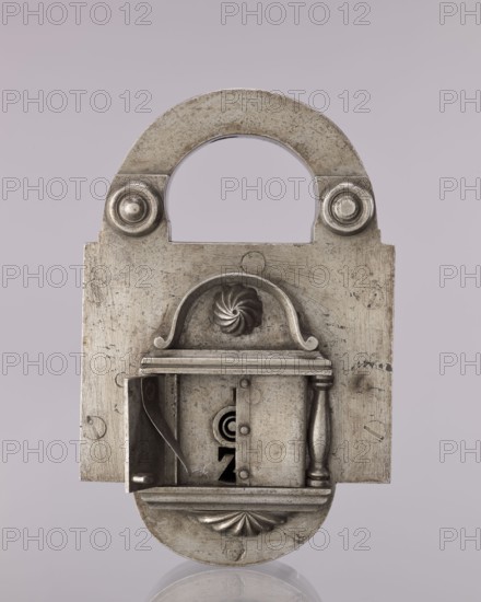Rectangular padlock with a bow, 19th century, Polesie, locksmithing; height 18.3 cm, width 11.7 cm, depth 5.4 cm, A close-up view of a metallic padlock with a rounded shackle and rectangular body, showing an ornate front plate with a small hinged compartment partially open revealing interior mechanisms; the metal surface appears aged with scratches and patina, and decorative elements include a shell-like motif, a carved spiral rosette, and a turned column detail. The visible colors are silver-gray metal tones with darker gray areas from tarnish and shadow, and a pale muted mauve-gray background.