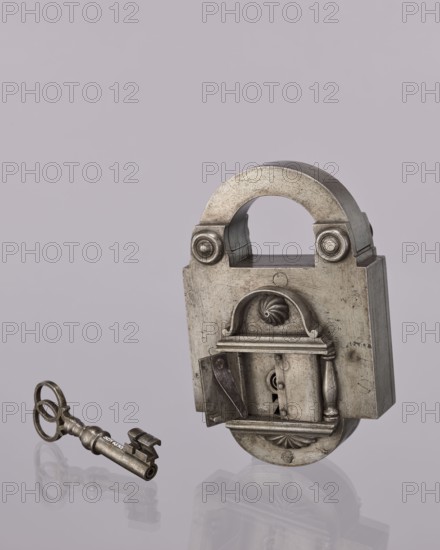 Padlock key, 19th century, Poland; locksmithing; height 10.3 cm, width 4 cm., A metallic padlock and a separate key sit against a pale lavender background, both showing a worn silver-gray surface with darker tarnished areas; the padlock has a rounded shackle and a rectangular body with decorative raised molding including curved and fluted motifs and a small hinged cover revealing an internal mechanism, while the key has an ornate bow with circular loops and a cylindrical shaft with notched bit, both objects casting soft reflections and faint shadows on the smooth surface below.