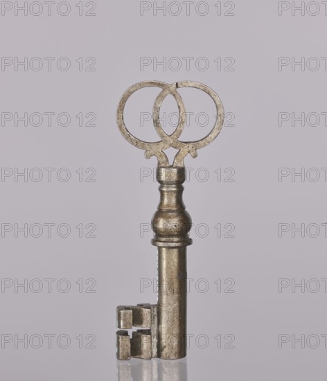 Padlock key, 19th century, Poland; locksmithing; height 10.3 cm, width 4 cm, A metallic key standing upright against a pale mauve background, featuring a rounded ornate bow formed by two interlocking circular shapes, a turned shaft with a bulbous midsection and a narrow neck, and a rectangular bit with a stepped cut; the key shows a worn, tarnished surface in shades of silver, gray, and muted brass, and a faint reflection is visible on the glossy surface beneath it.