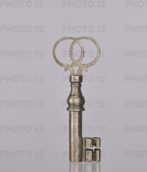 Padlock key, 19th century, Poland; locksmithing; height 10.3 cm, width 4 cm, A metal key stands upright against a pale gray background, with a rounded ornate bow featuring two overlapping circular loops and a textured, slightly tarnished surface showing silver and muted bronze tones; the shaft is cylindrical and smooth with a turned, bulbous section near the top displaying similar silver and bronze hues, and the bit at the base has a blocky, notched shape with worn edges, all reflected faintly on the glossy surface beneath.