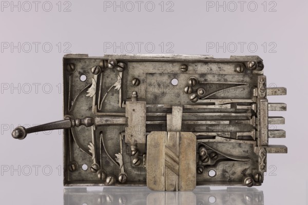 Gate lock. Double-bolt gate lock with Key No. 52. The lock is uncapped and has a pull handle. The entire mechanism shows very good, clean workmanship. Housed in a rectangular casing with five bolts: three central bolts slide back and forth, while the two outermost are scissor-type; when opened they lock the other bolts with side projections that engage cut-out recesses on the other bolts. Unlocking is achieved by lifting the lever at the end of the lock or by turning the key. Key register box closed with a rectangular plate with cut corners and a central strip with oblique cuts. Likely 17th/18th century, possibly Austria; locksmith work; height 17.5 cm, width 35 cm., A metal mechanical assembly with a rectangular frame and a lever extending from the left, showing a group of flat metal bars, curved spring-like components, and several rounded fasteners; the surface shows worn and aged metal tones including silver, gray, and darker tarnished patches, with a central block that has a warmer...