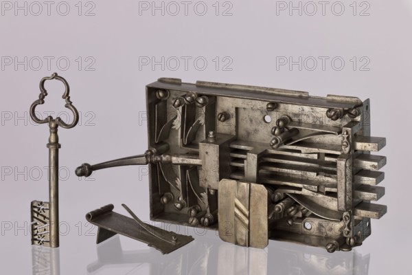 Gate lock. Double-bolt gate lock with key No. 52. Uncapped, with pull handle. Housed in a rectangular casing, with five bolts: three central bolts slide back and forth; the two outermost are scissor-type which, when opened, lock the other bolts via side projections engaging cut-out recesses. Unlocking achieved by lifting the lever at the end of the lock or by turning the key. Key register box closed with a rectangular plate with cut corners and a central strip with oblique cuts. Very good, clean workmanship; indicative of dedicated earlier craftsmen. 17th/18th century, possibly Austria; locksmith, Height 17.5 cm, width 35 cm., A metallic lock mechanism and key rest on a reflective surface against a pale background; the lock body is rectangular and open to reveal internal components including several flat bars, curved levers, screws, and a sliding block, all in a silvery metal with areas of darker tarnish and patina, while the detached key stands upright with an ornate bow and a long sh...