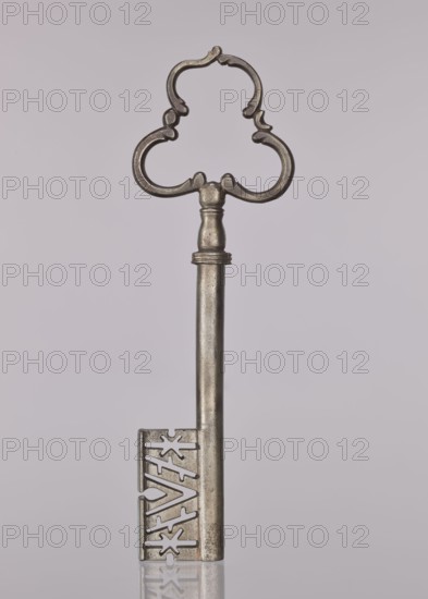 Gate key, locksmithing. Key with a tubular shaft, separated from the semi-ovoidal node by a flat cylinder; handle made of three C-sections. The blade widens downward, with a rib, diagonal notches and two stars. 17th/18th century, possibly Austria. Height 19.8 cm, width 6.2 cm, depth 1.3 cm., A single metal key stands upright against a pale gray background, with a decorative, ornate handle shaped like a symmetrical three-lobed loop showing smooth curves and small rounded protrusions, a tapered cylindrical shaft with a subtle band near the top, and a rectangular bit at the bottom featuring cut-out geometric shapes and starburst-like perforations; the key shows a metallic silver-gray color with slightly darker, aged patina in places and a faint reflection visible on the smooth surface beneath it.