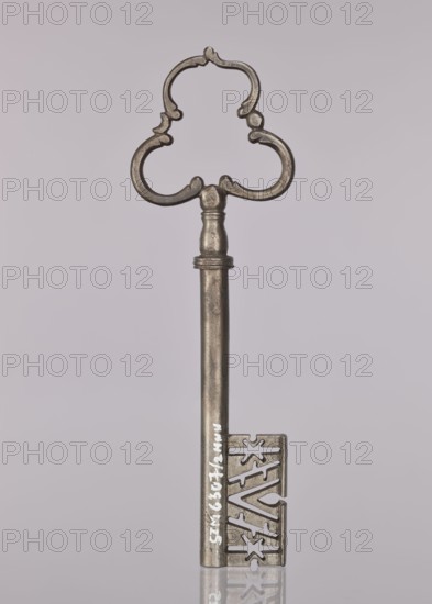 Gate key, locksmithing. Key with a tubular shaft, separated from the semi-ovoidal node by a flat cylinder; handle made of three C-sections. The blade widens downward, with a rib, diagonal notches and two stars. 17th/18th century, possibly Austria; height 19.8 cm, width 6.2 cm, depth 1.3 cm., A single ornate metal key is centered against a pale gray background, with a decorative clover-shaped bow featuring curved, stylized cutouts and a smooth cylindrical shaft leading to a rectangular bit that has intricate angular cut patterns; the key appears metallic with tones of silver and pewter, showing slight variations in darker and lighter gray, and it casts a faint reflection on the glossy surface below.