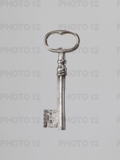 Iron key, locksmithing. Key with a tubular club-shaped shaft, with a feather widened at the end. Shaft joint with profiles, oval handle with bulges on the shorter axis. 1st half of the 19th century. Height 12.4 cm, width 4.5 cm, depth 1.2 cm. Maker, provenance: gift., A single metal key is centered against a plain light gray background; the key has an oval-shaped bow with a worn, slightly irregular outline, a short ornate neck with rounded ridges, a long cylindrical shaft that shows subtle scratches and patina, and a rectangular bit with several rectangular cutouts and notches. The visible colors are silver tones on the key and light gray in the background.