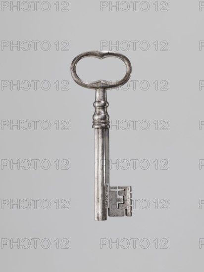 Iron key, locksmithing. Key with a tubular club-shaped shaft, with a feather widened at the end. Shaft joint with profiles, oval handle with bulges on the shorter axis. 1st half of the 19th century., and place. Height 12.4 cm, width 4.5 cm, depth 1.2 cm., A single metal key is centered against a plain light gray background; the key has a rounded, oval-shaped bow with a slightly irregular inner edge, a short neck with a few raised rings, a long cylindrical shaft showing areas of darker and lighter silver tones, and a rectangular bit with notches and a stepped profile. The visible colors are silver and light gray.