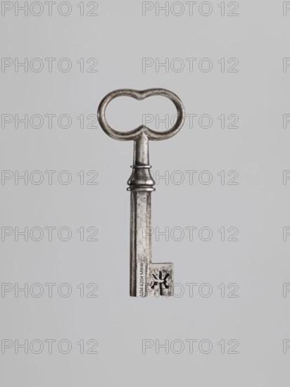 Key, locksmithing. Key with a tubular shaft with a Greek cross cross-section, with a feather slightly thickened toward the edge. Set in a knot with profiles and a vase element. Oval handle, concave at the knot and at the top. 19th century. Height 9.6 cm; width 4.1 cm; depth 1.3 cm., A single metal key is centered against a plain light gray background, showing a rounded bow with two connected loops, a short ornate neck with a ringed collar, and a long rectangular shaft ending in a bit with a cut-out floral-like shape; the metal appears aged with a dull silver-gray color and darker patina in crevices.