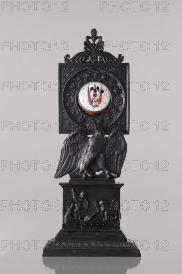 Watch stand with watch, Königlich-Preussische Eisengiesserei in Berlin fl. 1804–1874 ironworks or private foundries, watches, eagle iconographic, ca. 1830, Germany, Berlin, varia, height 31 cm, A dark ornamental clock with intricate carved details stands against a pale background; the clock features a rectangular carved panel topped with a decorative crest and a circular clock face set within a floral wreath, showing small colored painted elements on the dial. Below the panel sits a sculpted bird with spread wings perched on a rectangular pedestal that has a relief scene with winged figures and decorative motifs. The piece rests on a stepped base with carved borders. Visible colors include black on the carved structure, white and small areas of pink, purple, orange, and yellow on the clock dial, and a pale neutral background.