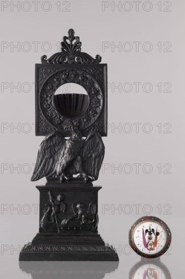 Watch stand with watch, Königlich-Preussische Eisengiesserei in Berlin fl. 1804–1874 ironworks or private foundries; watches; eagle iconography; ca. 1830; Berlin, Germany; varia; height 31 cm, A dark carved ornamental stand with a sculpted bird perched on a stepped base sits against a pale background, the stand featuring ornate floral and scrollwork details and a circular opening above the bird that frames a bowl-like recess; the base shows low-relief figures and decorative molding. To the right of the stand rests a round object with a reflective rim and a colorful center, showing hues of white, red, purple, gold, and black, and both items cast subtle reflections on the surface beneath them.
