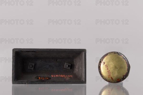 Watch stand with watch, Königlich-Preussische Eisengiesserei in Berlin fl. 1804–1874 ironworks or private foundries; watches; eagle iconographic; ca. 1830; Germany; Berlin; varia; height 31 cm, A rectangular dark metal box with a recessed interior sits beside a round metallic lid or disk; the box is matte black to dark gray with a rough texture and has two small square supports inside, while the round piece has a domed brass-colored center with a darker rim that shows patches of red and silver, and both objects rest on a reflective surface against a pale gray to light mauve background.