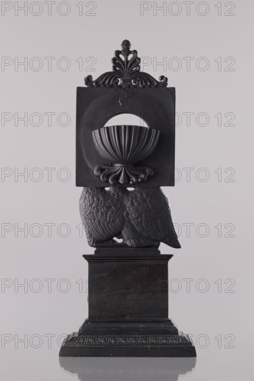 Watch stand with watch, Königlich-Preussische Eisengiesserei in Berlin fl. 1804–1874 ironworks or private foundries; watches; eagle iconography; ca. 1830; Germany; Berlin; varia; height 31 cm, A dark gray sculptural object stands against a pale gray background, featuring ornamental carved details and a square upper section with a semicircular cutout and a shell-like bowl shape, topped by a decorative crest with flowing scrollwork, supported below by a textured pair of wing-like forms with feather patterns, all resting on a stepped rectangular base with carved molding.