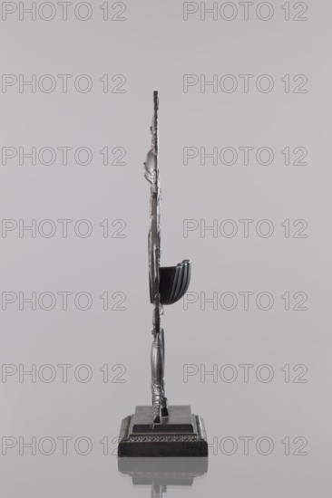 Watch stand, Königlich-Preussische Eisengiesserei in Berlin fl. 1804–1874,  ironworks author or private foundries, watches, panoply ornament, ca. 1830, Berlin, varia, height 26.5 cm, width 13.5 cm, depth 6.5 cm, A thin vertical metallic sculpture shown from its narrow side, standing on a decorative rectangular base with a reflective surface beneath; the sculpture and base are primarily silver and dark gray with subtle highlights, and the background is a smooth pale gray.
