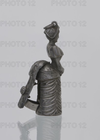 Cigar cutter, tobacco accessory, women's fashion; cigar cutter in the form of a full-plastic figurine of a lady in an outfit from the 1880s — a dress with a bustle — which contains the mechanism of the device that, when pressed, activates a guillotine. Unmarked, late 19th century, accessory; height 5.6 cm, width 1.2 cm., A small metallic figurine of a woman seen in profile standing on a reflective surface, rendered in dark gray and silver tones; she wears a fitted bodice with short sleeves and a long, draped skirt with layered, gathered folds, and a long scarf or shawl that hangs from her shoulder and wraps behind her, creating a looped shape; her hair appears styled and is topped with a small ring or loop element; the surface shows varied textures with smooth and slightly rough areas and subtle highlights and shadows in gray and silver.