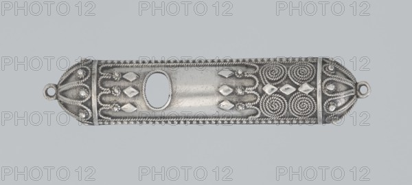 Mezuzah, 19th century, Israel; Judaica; height 0.6 cm, width 6.6 cm, depth 1.3 cm, weight 9 g., A long horizontal metallic object with decorative raised patterns occupies the center of the image; it is primarily silver with darker gray areas accentuating shadows and recessed details. The piece has rounded ends each featuring a small circular loop, and sections of twisted rope-like ridges run along the edges and form spiral motifs near one end. There are several raised diamond-shaped elements and small rounded beads arranged in patterns across the surface, and an oval cutout appears in the central area surrounded by a finely textured border. The background is a uniform pale gray-blue.