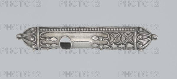 Mezuzah, creator unknown, 19th century, Israel; Judaica; height 0.6 cm, width 6.6 cm, depth 1.3 cm, weight 9 g., A horizontal cylindrical metal object with decorative raised patterns and a central oval opening, shown against a light gray background. The object appears silver in color with darker gray areas in recessed details. Both rounded ends feature looped attachments and braided rope-like motifs with small raised bead elements. Along the length there are twisted spiral patterns, diamond-shaped raised pieces, beaded borders, and rope-like twisted lines creating symmetrical ornamental designs. The surface shows polished highlights and subtle scratches, and the overall color palette includes silver and shades of gray.