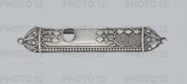 Mezuzah, 19th century, Israel, Judaica; height 0.6 cm, width 6.6 cm, depth 1.3 cm, weight 9 g., A long, narrow metallic object with a silvery-gray appearance rests against a pale gray background; its surface is decorated with ornate raised patterns including twisted rope-like spirals, rounded bead rows, teardrop shapes, and repeating diamond-like raised elements, with smooth polished central areas that show fine surface scratches and an oblong cutout near the middle; both rounded ends feature looped rings and symmetrical rope and bead embellishments, and the overall color palette includes silvery-gray and darker gray tones for shadows and recessed details.