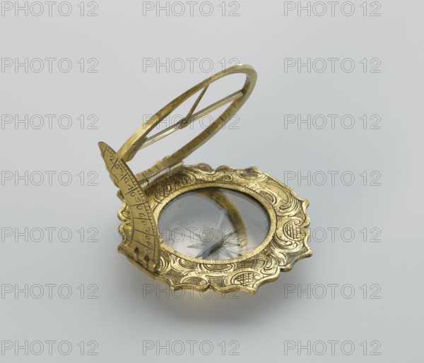 Equatorial sundial by Andreas Vogler ca. 1730–1800. Augsburg-type circular sundial with a wavy edge. Inside the case: a compass with a hinged hour circle and an arched hinged arm marked with geographical latitudes. Engraved decoration on the rim with C-shaped sections and stylized acanthus leaves. Engraved on the underside of the compass case: Eleva. Poli. / Corfu 39 Lyon 45 /' Berlinn 53 Cadiv 36 / Lisabon 39 Paris 48 / Colln 521 Reval 61. Dated to the 3rd quarter of the 18th century, Augsburg, Germany. Measuring instrument sundial., A decorative metallic object with a scalloped, ornate rim in gold tone sits on a pale gray surface; the object has a circular glass-covered center reflecting muted gray and some darker tones, and an upright arched element in the same gold tone attached to the rim, with a gold oval ring mounted above it.