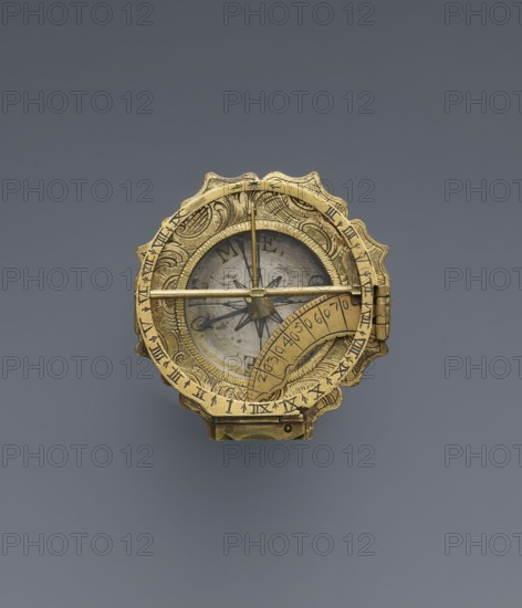 Equatorial sundial by Andreas Vogler ca. 1730–1800. Augsburg-type circular sundial with a wavy edge. Inside the case: a compass with a hinged hour circle and an arched hinged arm marked with geographical latitudes. Engraved decoration on the rim with C-shaped sections and stylized acanthus leaves. Underside of the compass case engraved: Eleva. Poli. / Corfu 39 Lyon 45 /' Berlinn 53 Cadiv 36 / Lisabon 39 Paris 48 / Colln 521 Reval 61. Dated to the 3rd quarter of the 18th century, Germany Augsburg. Measuring instrument., A small circular metallic object with ornate scalloped edges in gold color sits against a smooth gray background; the object has intricate engraved patterns and a hinged clasp, a glass-covered circular face showing a star-shaped design and a central needle, a thin horizontal bar crossing the face, and a curved engraved plate partially covering one side.
