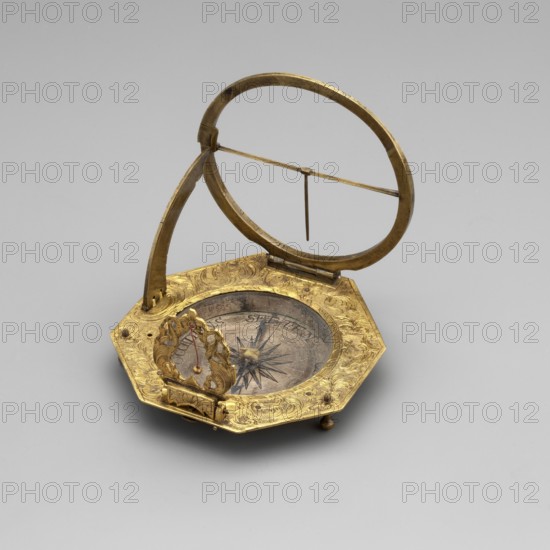 Equatorial sundial, mid-18th century, Augsburg, Germany. Sundial with compass resting on legs, in a frame on an octagonal base, decorated with an engraved plant-floral ornament flowers, with a compass at the center; movable rim with Roman numerals. Timepiece; height 7.7 cm, width 7.6 cm., A small octagonal metal object with a patinated golden surface rests on a neutral light-gray background; it has an ornate engraved rim with swirling floral-like patterns and a circular recessed center containing a round element with a star-shaped design and a dark radial motif. A decorative vertical metal frame rises from one side and supports an open oval ring above the center, with a thin horizontal bar crossing the ring and a short vertical pointer hanging from the bar. At the front of the object a sculpted ornamental piece with curved leafy forms and a small suspended bead-like element stands upright above the recessed center. The object shows areas of darker aged metal and brighter golden highlig...