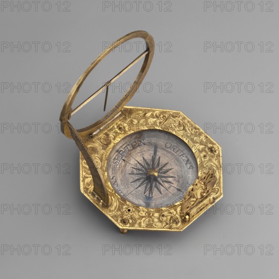 Equatorial sundial, mid-18th century, Augsburg, Germany. Sundial with compass resting on legs, in a frame on an octagonal base, decorated with engraved plant and floral ornament flowers, with a central compass and a movable rim with Roman numerals. Timepiece; height 7.7 cm, width 7.6 cm., An octagonal metallic object with a hinged circular ring rising from one side and a thin bar across the ring, all in a gold tone, sits on a flat gray background; the central circular area features a radial starburst pattern in darker gray and black tones surrounded by a weathered bluish-gray disk, and the gold-toned rim around the disk is decorated with ornate floral and scrollwork engravings; a small ornate component in gold tone projects from one edge, and a few tiny rounded feet in gold tone lift the piece slightly off the surface.