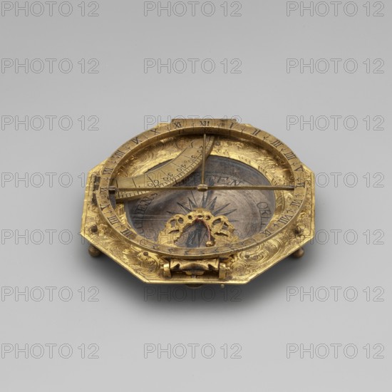 Equatorial sundial, mid-18th century, Augsburg, Germany; timepiece. Sundial with compass resting on legs in a frame on an octagonal base, decorated with engraved plant and floral ornament centered around a compass; movable rim with Roman numerals. Height 7.7 cm, width 7.6 cm., A small octagonal metal instrument sits on a plain light gray surface, primarily gold in color with darker brown and black tones in its central circular area; the metal has ornate engraved decorative patterns and a raised rim around the central cavity, with a curved graduated arc and thin metal crossbars spanning the interior; a decorative scalloped element with small raised knobs occupies one side of the central area, and a starburst-like motif is visible beneath the crossbars; the piece rests on small rounded feet and shows areas of polished shine and subtle wear.