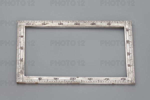 Ruler with scale. Rectangular frame with scale made of silver-plated brass, 18th century, Western Europe. Measuring instrument. Height 13.7 cm; width 3.2 cm., A rectangular metallic frame with a polished silver appearance rests on a smooth gray background; the frame has an inner rectangular opening and a narrow border decorated with a series of small notches and engraved linear markings around its perimeter, showing subtle variations in the silver surface with areas that appear slightly darker or tarnished.