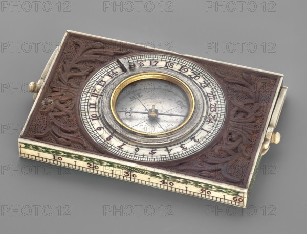 Compass, measuring instrument, ca. mid-18th century, Western Europe. Compass with a bone scale around the dial with cardinal directions and a hand behind the glass, in a wooden frame decorated with relief acanthus, with a bone lining decorated with Regency ornamentation. Height 13.3 cm; width 8 cm., A rectangular object with a carved dark brown surface featuring ornate leaf-like patterns surrounds a central circular inset containing a metallic disc and a glass-covered needle; the circular rim has metallic silver and gold tones with an inner gold ring and an outer silver ring, and a few small dark metal components are visible on the rim. The sides and edges of the object are light cream or ivory with decorative green bands and small dark markings, and there are small rounded cream-colored knobs on the sides. The background is a smooth neutral gray.