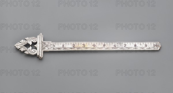 Ruler with a handle composed of interconnected acanthus twigs forming a triangle. Measuring instrument, ca. mid-18th century, Western Europe. Materials/ornament: acanthus. Height 14.7 cm, width 3.2 cm., A decorative metal ruler with an ornate filigree handle in silver and light gray tones rests horizontally against a smooth medium gray background; the ruler's long straight body shows a slightly worn metallic surface with darker gray and faint warm discoloration, and the handle features intricate leaf-like scrollwork in silver and gray.