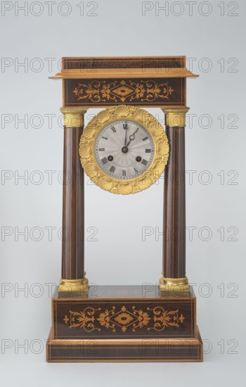 Mantel clock by Alexandre Pertrand fl. ca. 1840. Four-column portico form on cuboid base and counter-base; boxwood inlays on front of cornice and base with rhombus rosette and tendril on both stilts; gilded bronze bases and capitals. Suspended movement with silver dial with Roman numerals and two keyholes hangs between the columns. Gilded bronze frame with shells surrounds the clock. Oak construction with rosewood facing and light wood insets. Circa 1840, Paris, France. Timepiece; height 47 cm, width 21 cm., A decorative vertical mantel clock with dark brown wood, gold, and light gray tones, featuring a rectangular dark brown base with inlaid lighter brown floral patterns and gold trim, two dark brown fluted columns with gold capitals and bases rising from the base to support a rectangular dark brown top shelf that also has lighter brown floral inlay and gold edging, and a central round clock face suspended between the columns with a gold ornate circular frame and a light gray inner di...