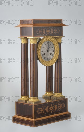 Mantel clock by Alexandre Pertrand fl. ca. 1840. Mantel clock in the form of a four-column portico on a cuboid base and counter-base; boxwood inlays on the front of the cornice and base, with a rhombus rosette and a tendril on both stylons; gilded bronze bases and capitals. A suspended movement with a silver dial with Roman numerals and two keyholes hangs between the columns. A gilded bronze frame with shells surrounds the clock. Oak construction with rosewood facing and light wood insets. Ca. 1840, Paris, France. Timepiece; height 47 cm, width 21 cm., The image shows an ornate pedestal clock with a rectangular wooden top and base connected by a group of four tall cylindrical columns. The wood has dark brown tones with lighter brown inlay and decorative floral marquetry on the top and base. The columns are topped and footed with gilded metal collars and mounts in a yellow-gold color. A round clock face is suspended between the columns, surrounded by an elaborate yellow-gold ornamental ...